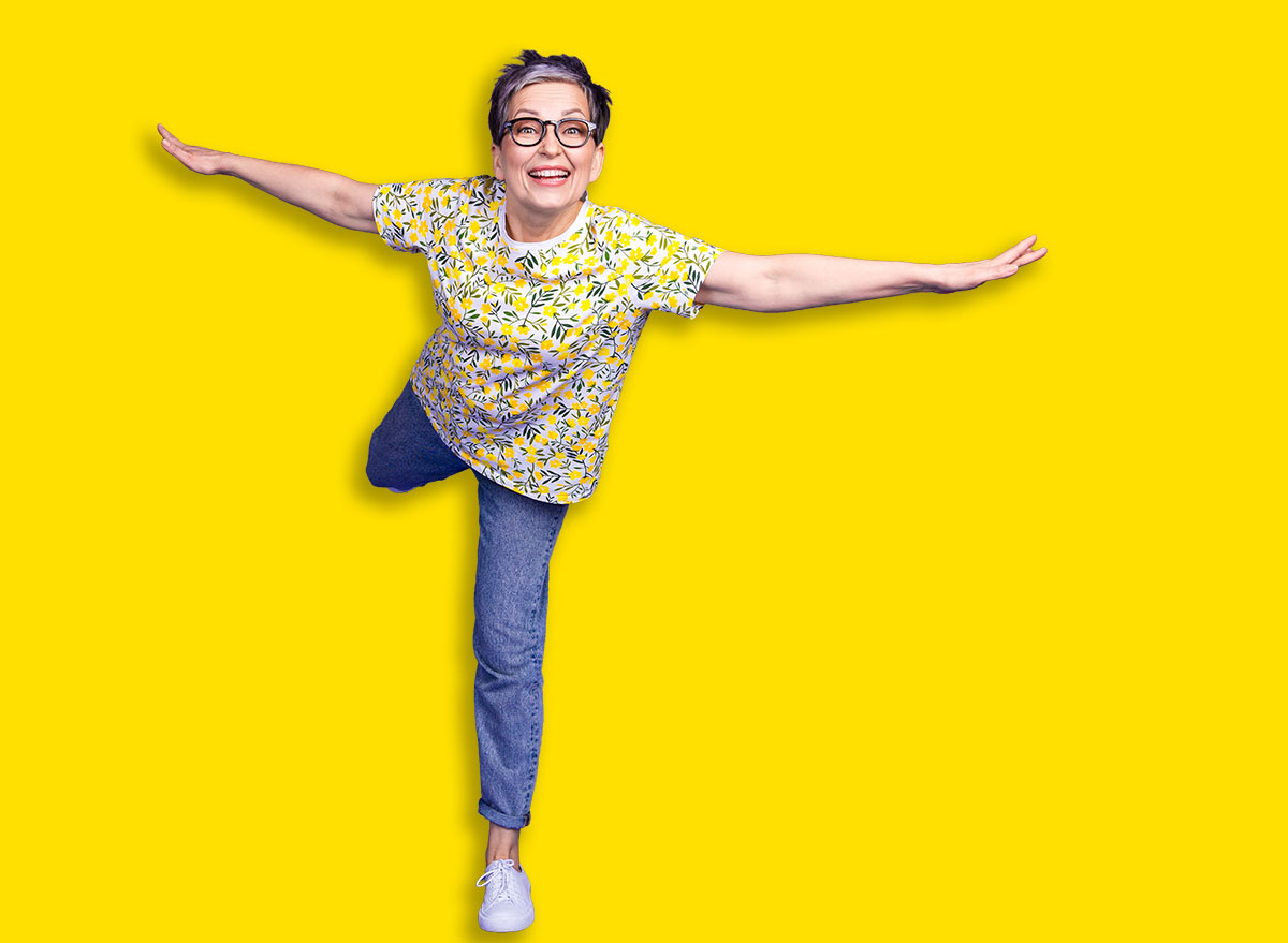 Happy senior woman balancing playfully on vibrant purple background, wearing casual yellow flower print shirt with jeans
