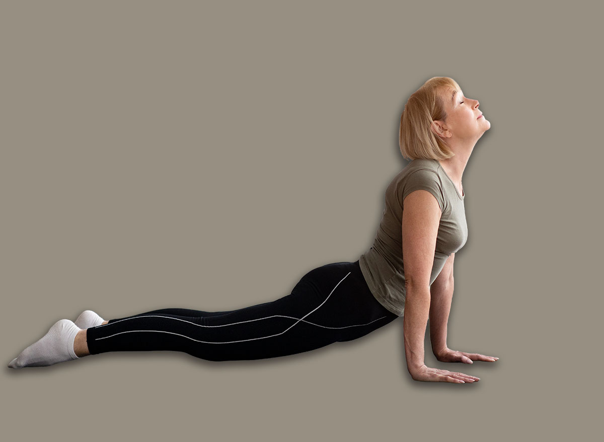 Full length of fit senior woman doing cobra pose on yoga mat in living room, empty space. Side view of athletic mature lady doing flexibility exercises, working out during covid-19 isolation