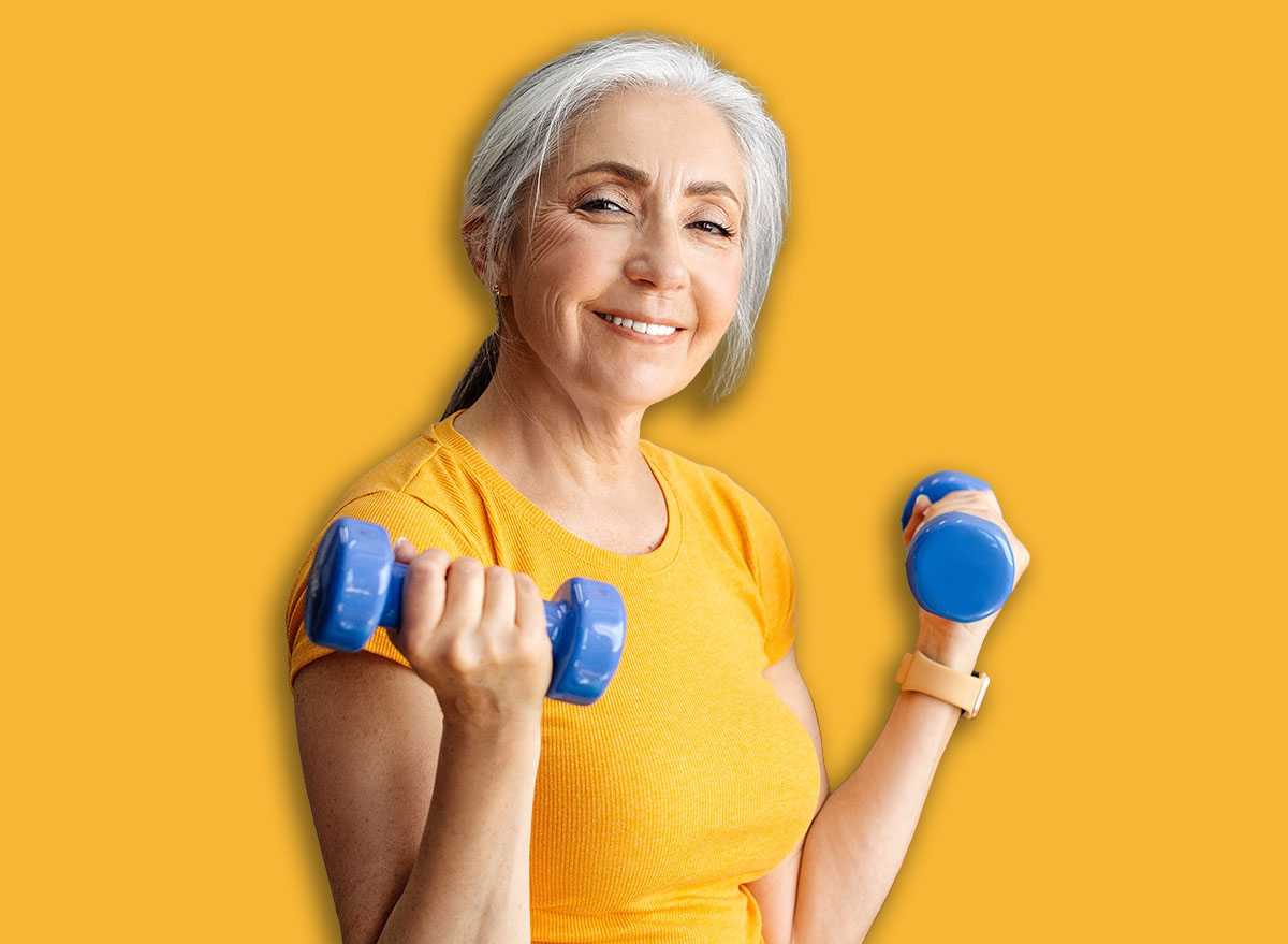 Happy senior woman doing dumbbell workout at home, working on arms strength, looking at camera and smiling. Fit aged lady lifting fitness dumbells up over living room interior