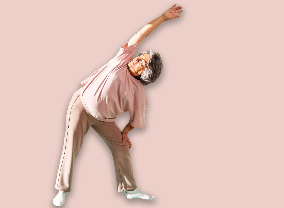 A senior woman performs a side stretch at home, standing on a yoga mat in a sunny living room. She enjoys her daily exercise routine surrounded by plants and fitness equipment.