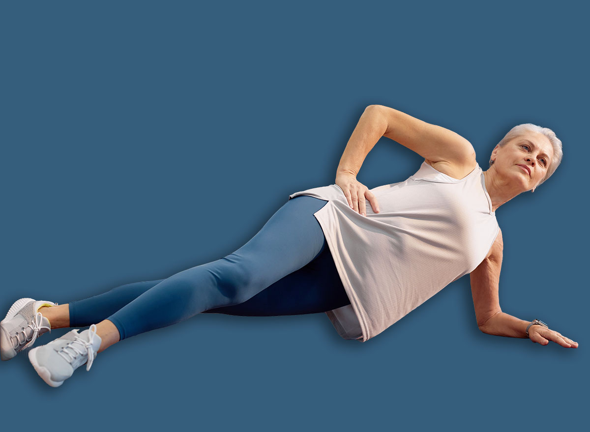 Senior Caucasian woman exercising on yoga mat performing side plank in modern gym, supporting body with arm and looking focused, fitness equipment visible in background