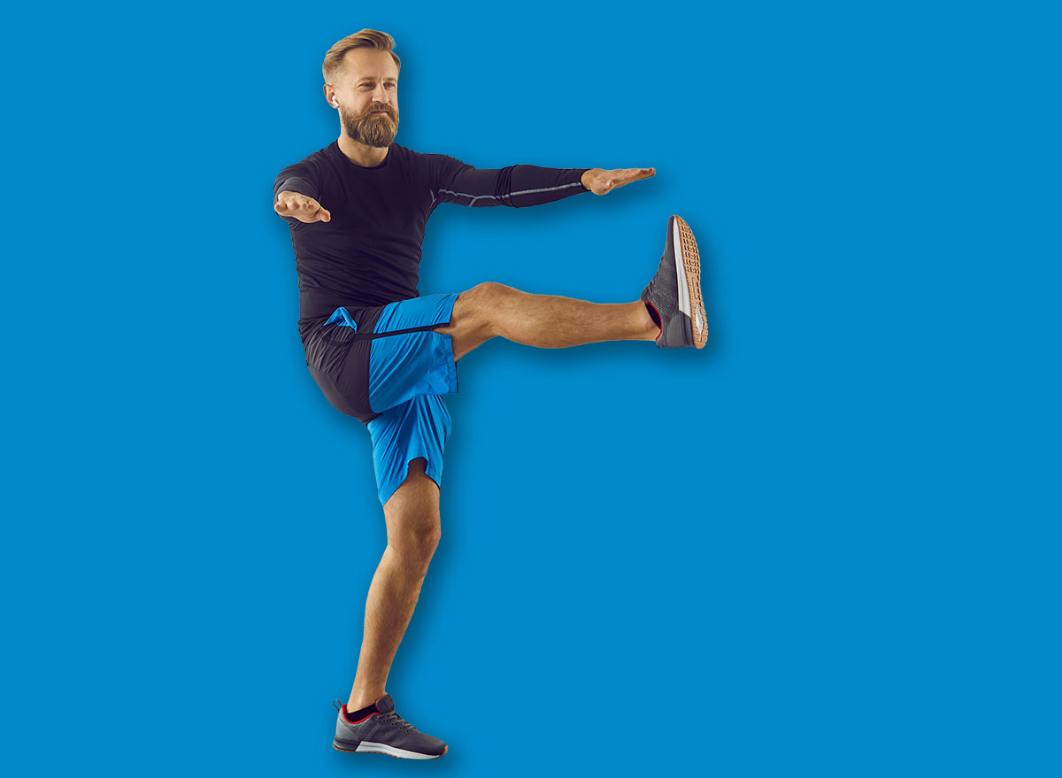Fit man having active workout at home. Handsome strong athlete following individual fitness program doing leg swing exercise on sports mat and using modern mobile phone app to track physical activity