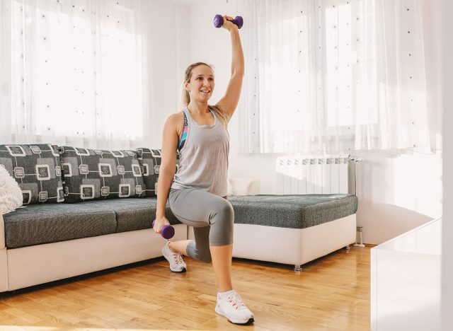 Smiling muscular sportswoman in shape doing lunges while holding dumbbells.