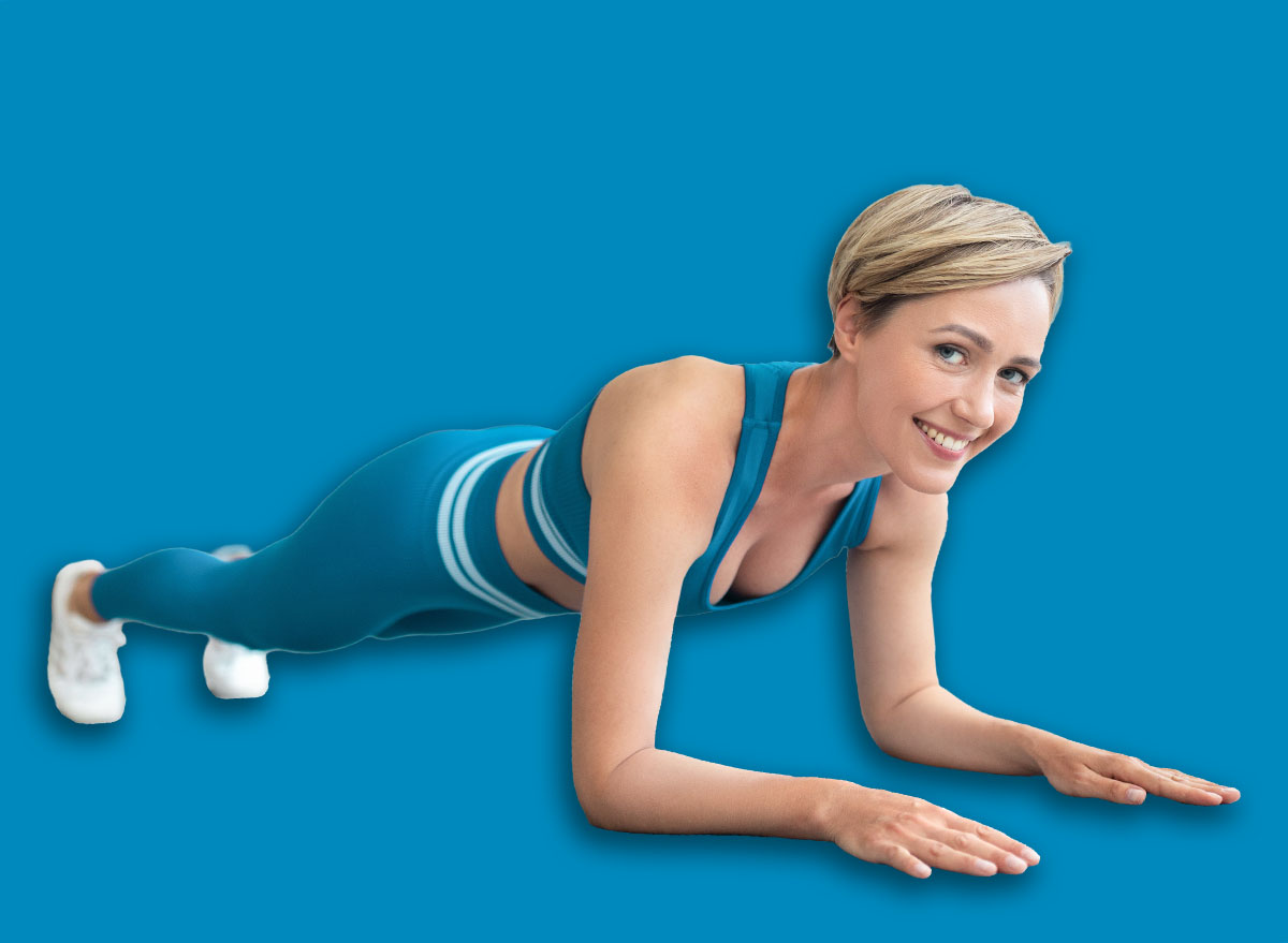 Healthy Lifestyle Concept. Smiling Fit Lady Standing In Elbow Plank Position On Yoga Mat At Home In Living Room Looking At Camera. Cheerful Slim Female In Sportswear Training Core And Abs Muscles