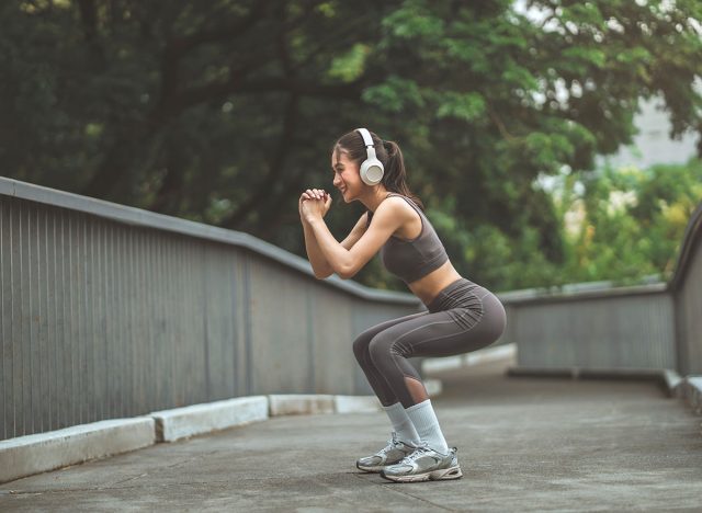 Happy fitness Asian woman wearing wireless headphones doing squat exercise workout at public park – active healthy lifestyle, outdoor training, warm-up routine and body care in urban green space