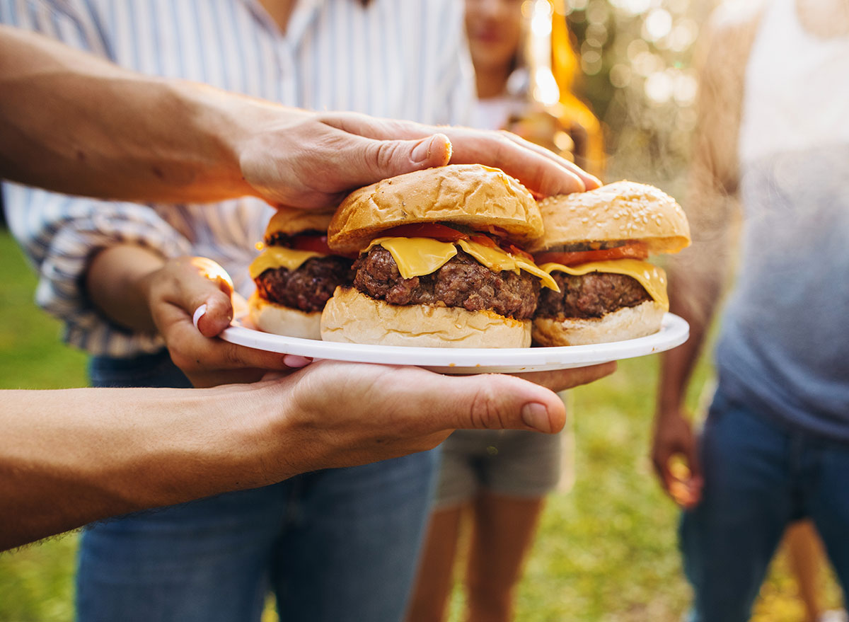 Delicious. Image of tasty homemade burger with melting cheese, tomatoes and grilled meat. Group of friends having nice outdoor picnic, making food, drinking beer and having fun.