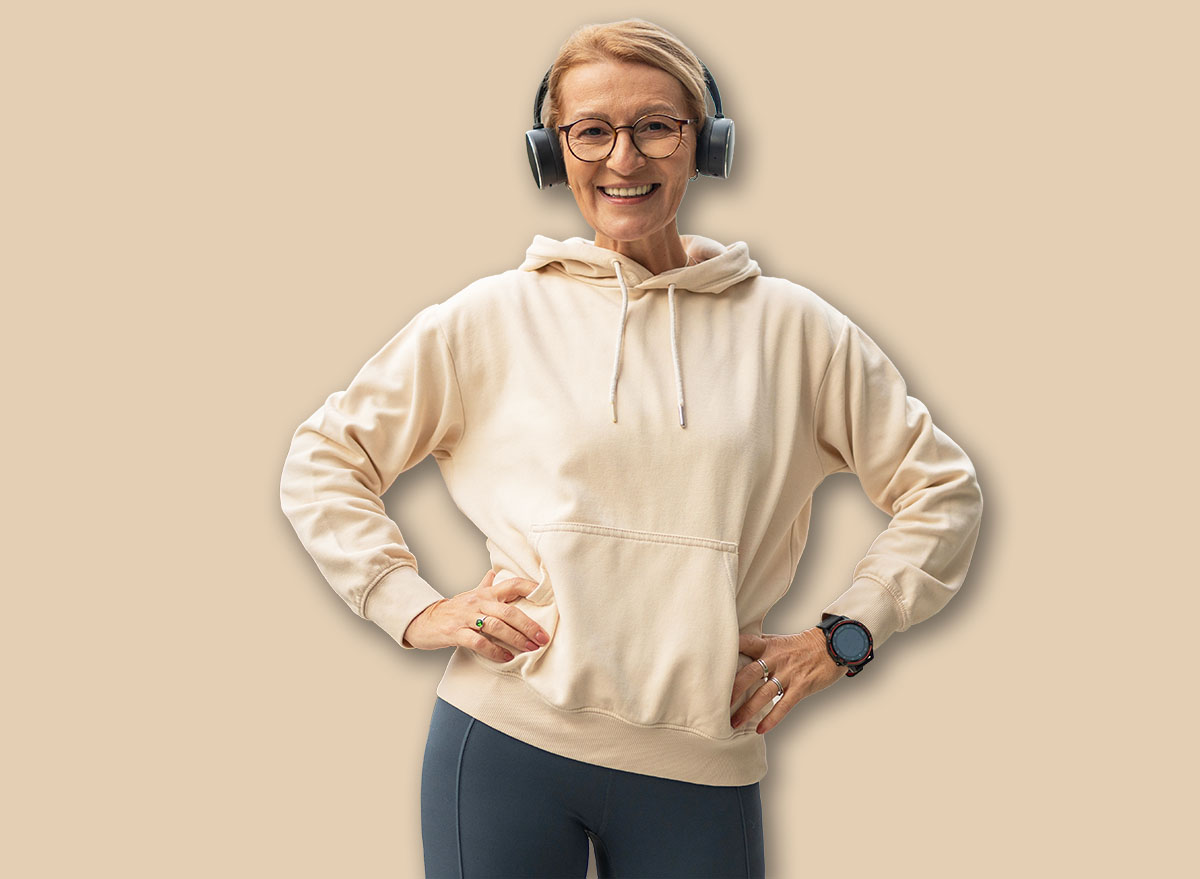 Mature woman wearing headphones and sportswear, standing confidently with hands on hips, smiling at the camera, embodying an active and healthy lifestyle in a modern city environment