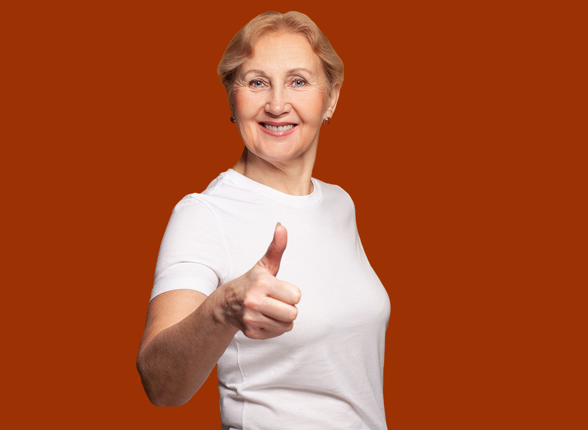 Happy senior woman in white t-shirt showing thumb up on bright yellow studio wall banner background