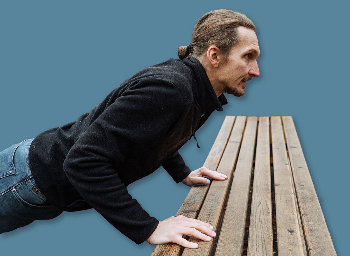 athletic man doing incline pushups in park, person executing incline pushups with park equipment nearby, individual engaging in incline pushups on wooden bench at outdoor fitness park