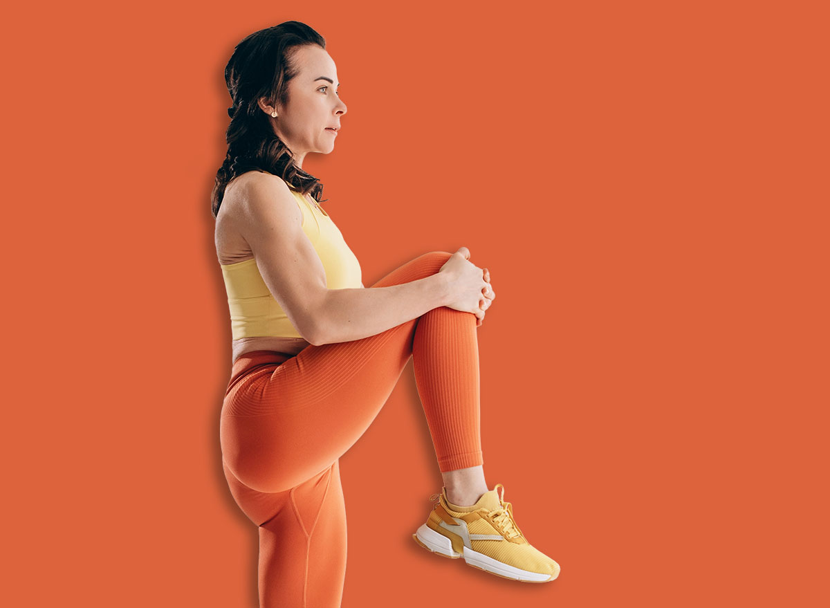 Athletic woman doing standing knee to chest stretch during warm up workout in bright modern exercise studio