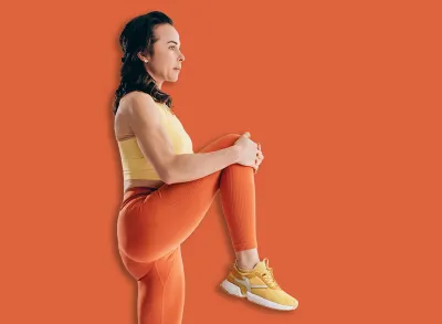 Athletic woman doing standing knee to chest stretch during warm up workout in bright modern exercise studio