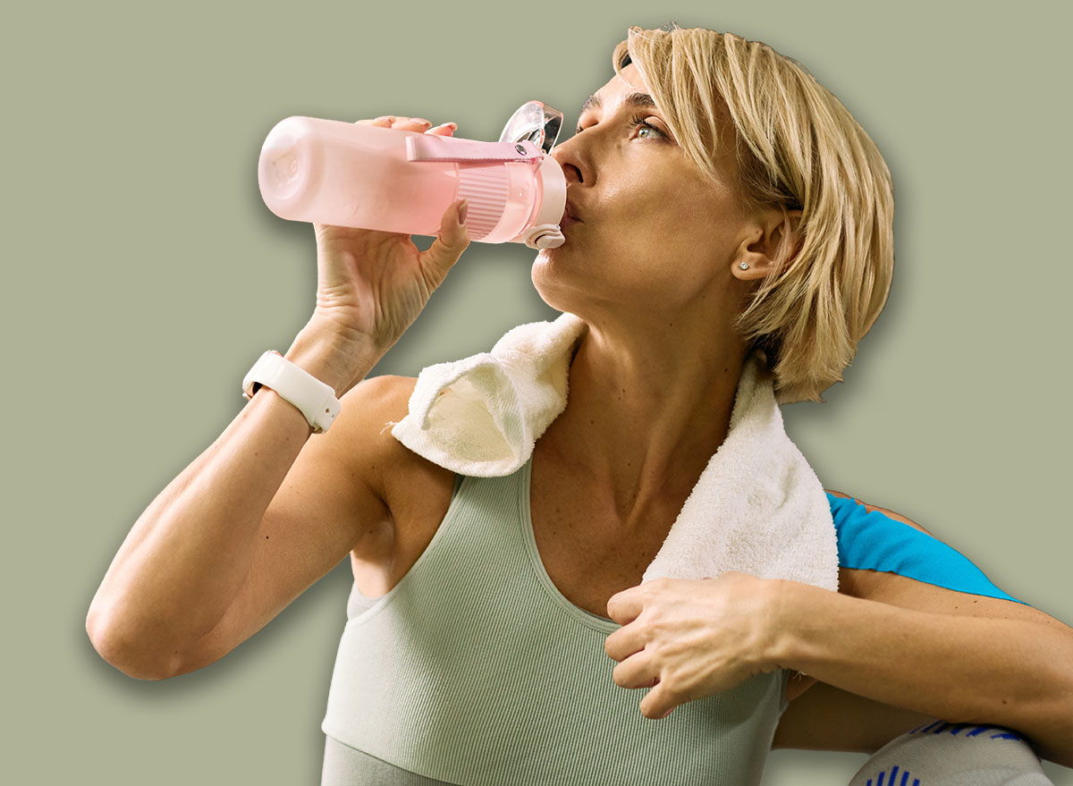 Caucasian middle aged woman hydrating with sports bottle during physical therapy session, towel draped over shoulder, engaging in rehabilitation exercise in gym setting