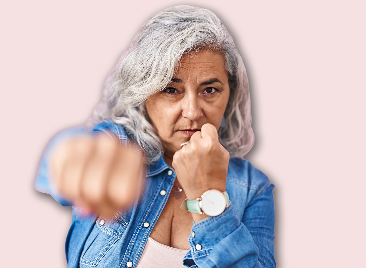 Middle age woman with grey hair standing over pink background punching fist to fight, aggressive and angry attack, threat and violence