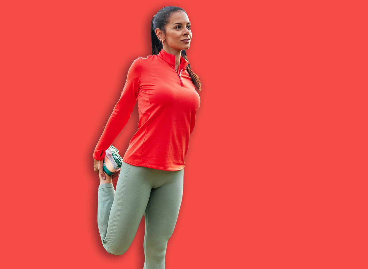 Brunette caucasian woman in running shoes stretching her quad muscle wearing a bright pink long-sleeve top and light green leggings outdoors by a lake.