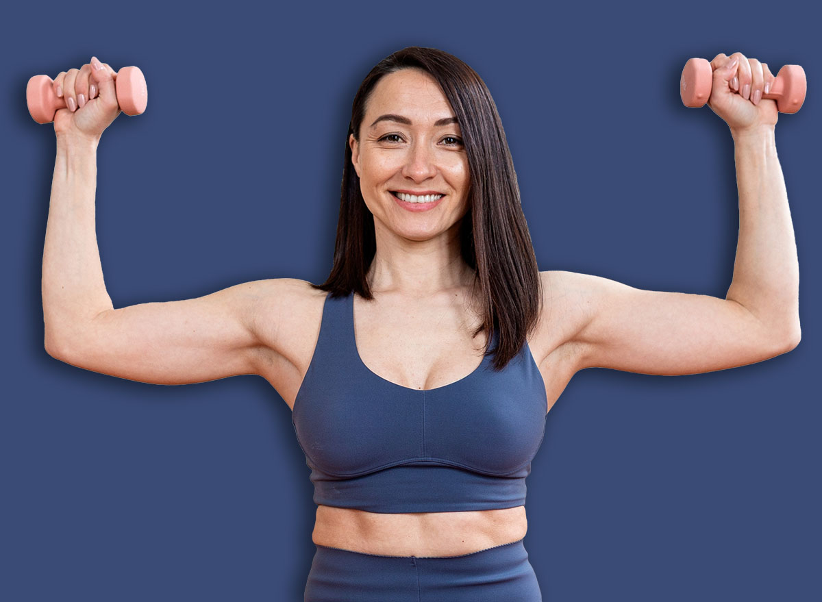 Smiling Asian woman performing a dumbbell overhead shoulder press while kneeling on a yoga mat. Concept of upper body strength, home weight training, functional fitness, and muscle toning.