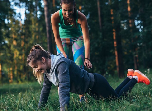 Sporty young female trainer showing a girl how to do knee push-ups while training outdoors in summer