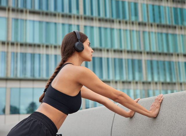 Sporty slim woman does push up exercises leans on concrrete wall wears cropped top stereo headphones on ears has pigtail poses outside has workout in open air. Motivated sportswoman in activewear