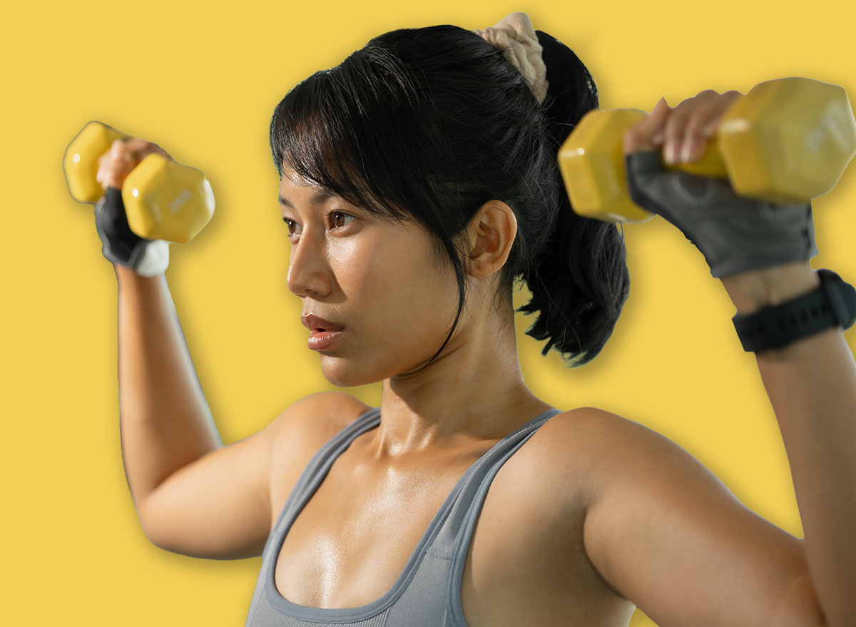 Asian woman in gray sportswear presses yellow dumbbells overhead during a shoulder workout in a bright gym.