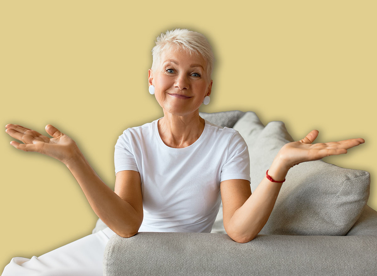 Wise senior female with calm smile on couch throwing up hands in despair. Something went wrong, fail of plans, ruined day. It is what it is, acceptance, go with flow concept
