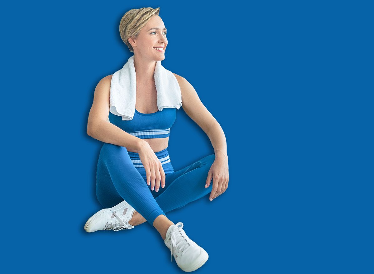 Domestic Train Concept. Smiling woman exercising at home, taking break, resting and thinking, looking away at window. Middle-aged lady sitting on yoga mat in living room with towel on shoulders