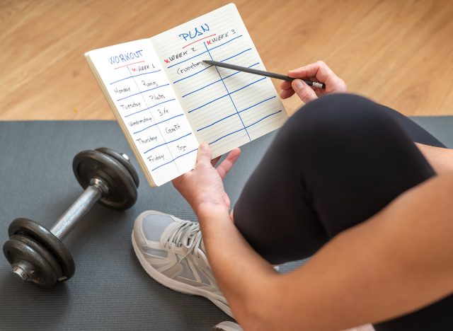 Active Female Athlete Preparing Workout Routine On Notepad While Sitting On Yoga Mat With Dumbbells, Promoting A Healthy And Active Lifestyle, Focused On Fitness Goals, Morning Exercise Motivation