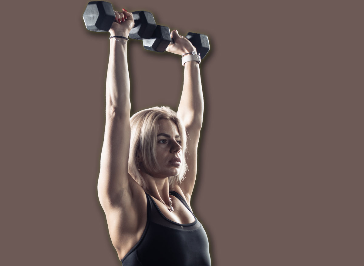 Athletic woman in sportswear trains shoulder muscles with heavy dumbbells in her hands in gym. Healthy lifestyle, fitness and bodybuilding concept.
