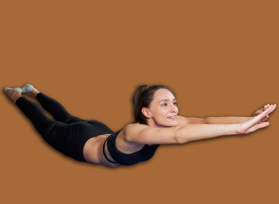 A smiling sporty girl in a black workout tight suit is doing the classical boat exercise for the back. A coach in a superman pose conducting a remote online fitness class on the blue yoga mat at home.
