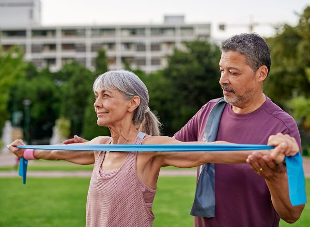 Senior multiethnic couple using a resistance band during outdoor exercise. Multiethnic man helping beautiful mature woman withstretching exercise while using a blue band. Older adults doing fitness.