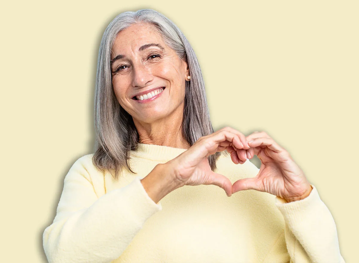 middle age gray hair woman smiling and feeling happy, cute, romantic and in love, making heart shape with both hands