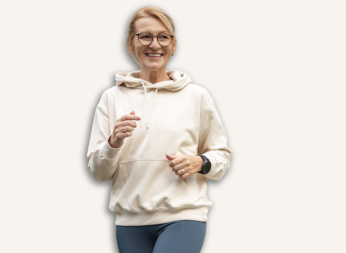 Senior woman in activewear jogging through a modern urban neighborhood, smiling and energized while staying fit outdoors with smartwatch and casual hoodie on a sunny path
