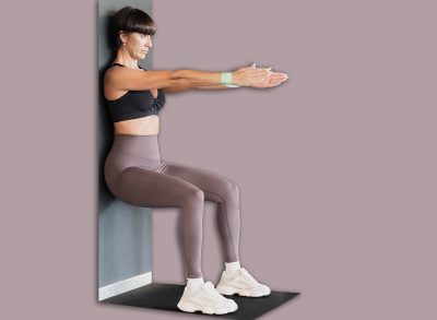 Female personal trainer coaches young woman through wall sit exercise beside foam roller in bright contemporary gym, offering hands-on support, posture correction, motivation, and focused lower body