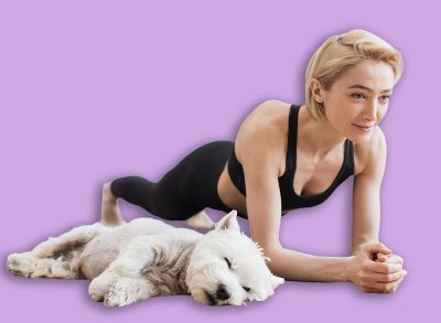 Peaceful blonde lady in black activewear doing yoga practice while tired Westie resting beside on floor of kitchen. Athletic yogini holding Forearm Plank Pose during yoga session with pet at home.