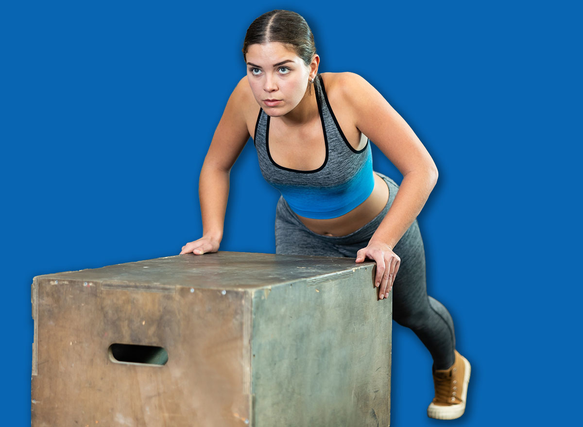 Motivated sporty young girl performing incline push-ups on plyometric box, focused on chest, shoulders, triceps, and abs training during intense group workout at gym