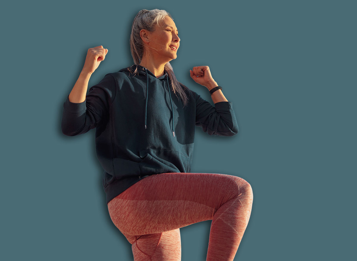 Active female in pink leggins and blue hoodie exercising , lifting one knee up and looking ahead on sunny day