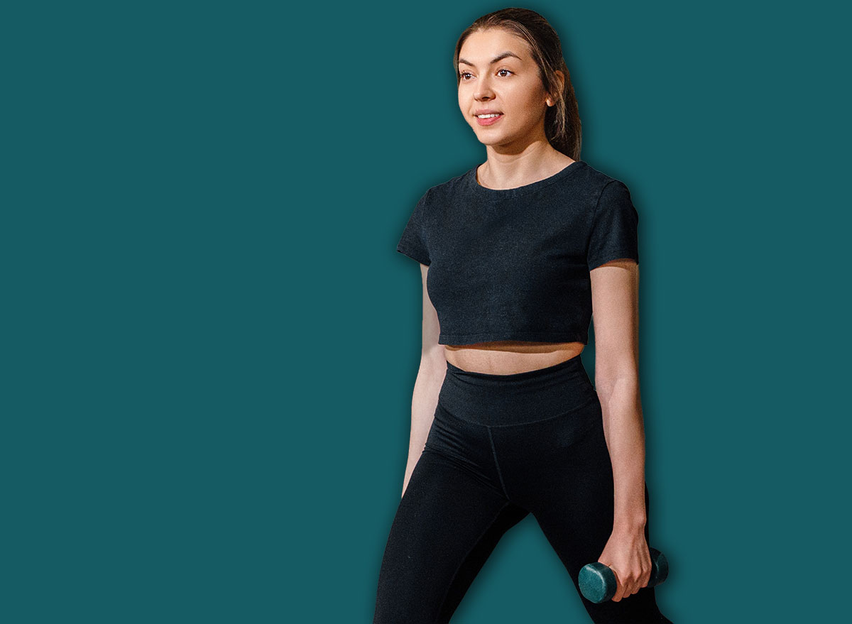 Pretty woman completing forward lunges with stable strong posture. Portrait of female model carrying dumbbells in modern gym, active training concept.