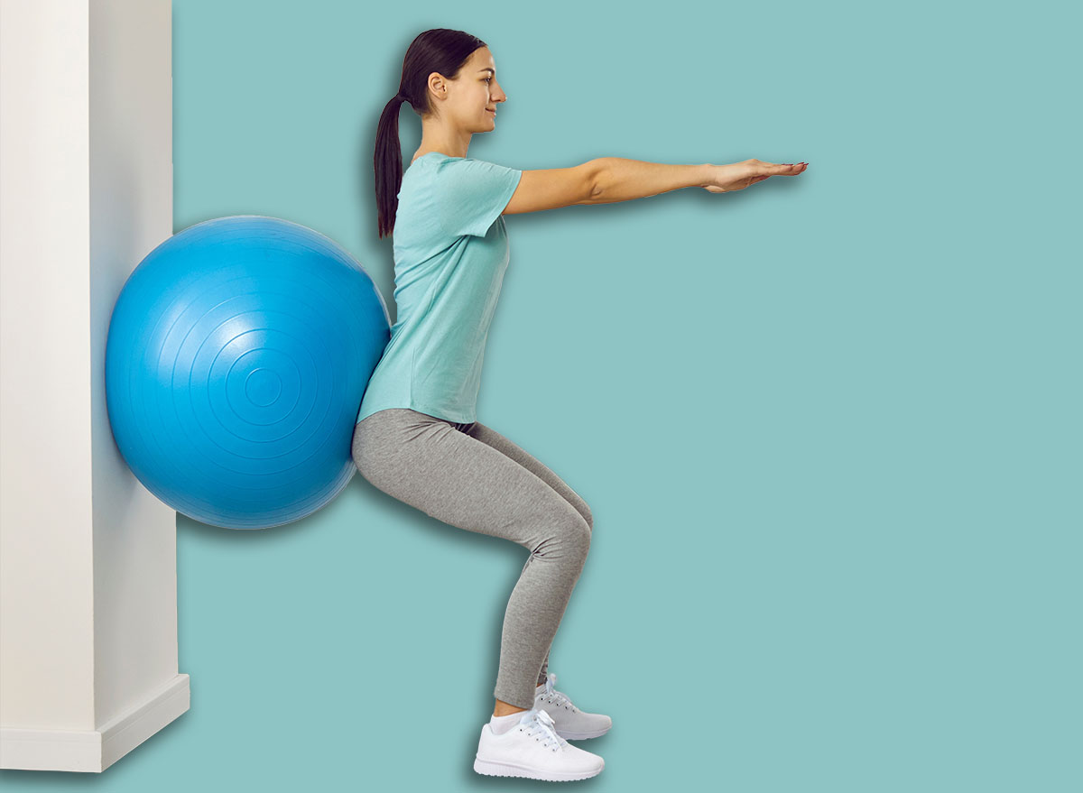 Fitness trainer helping young female patient do wall squats with blue fit ball to get rid of backache and regain spinal health. Woman doing back exercise in physio room of modern clinic