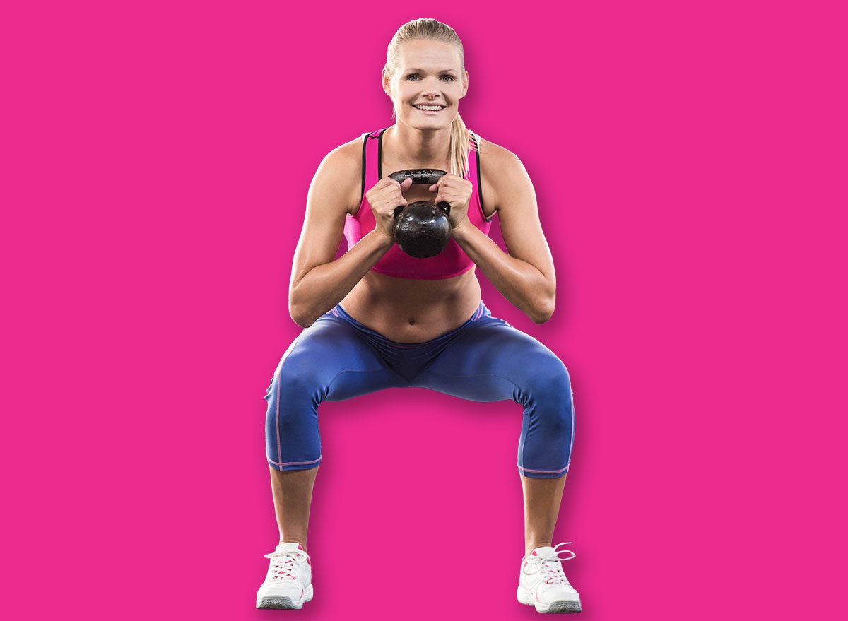 Woman holding kettlebell at chest while squatting in fitness studio with colored weight plates. Gym, fitness, strength, workout, training, exercise, wellness