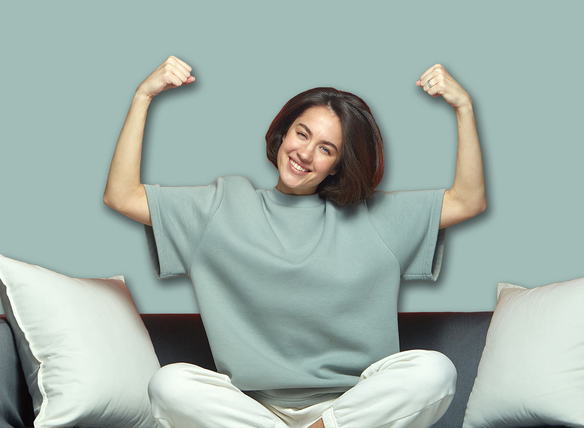 Successful young girl showing power raised arms, demonstrate biceps muscles proud of achievements, sitting on sofa