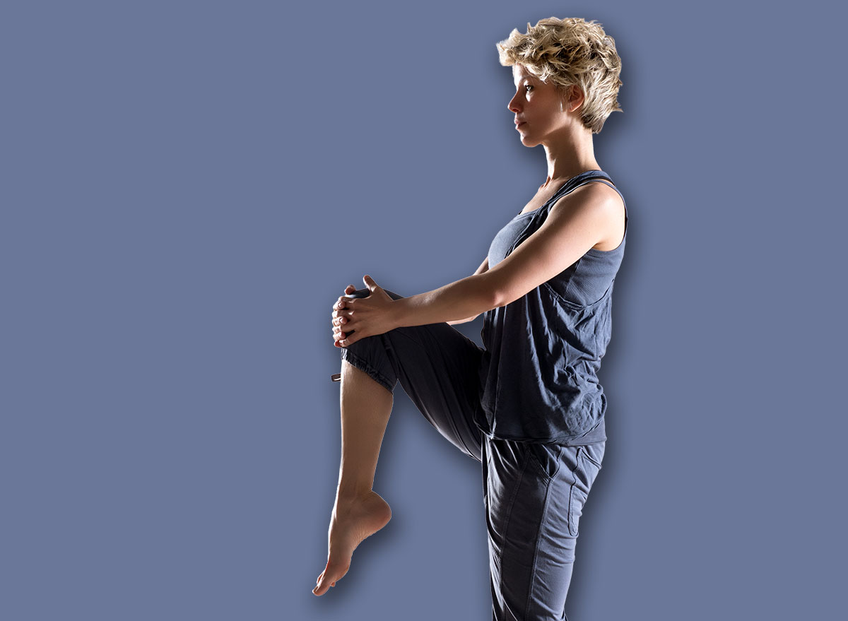 Side view on balanced woman standing tall on hardwood floor with one leg while holding knee up
