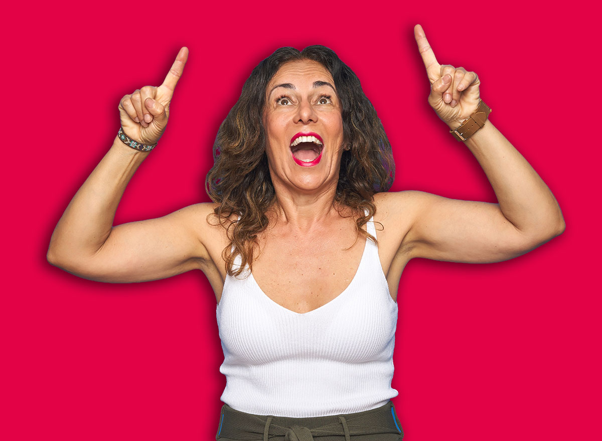 Middle age senior woman with curly hair standing over blue isolated background smiling amazed and surprised and pointing up with fingers and raised arms.