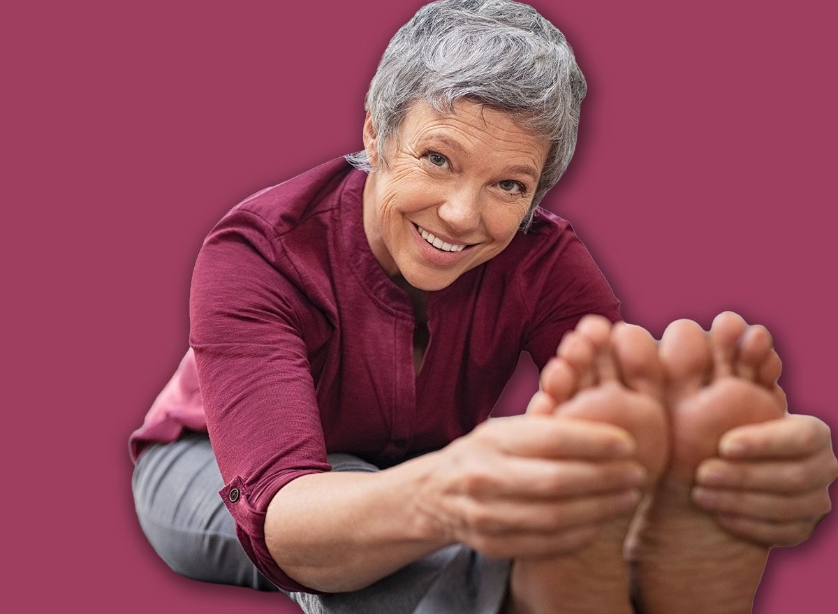 Closeup of senior woman stretching to touch toes while sitting on yoga mat. Portrait of mature woman doing her stretches at home while looking at camera. Happy healhty lady doing yoga exercises.