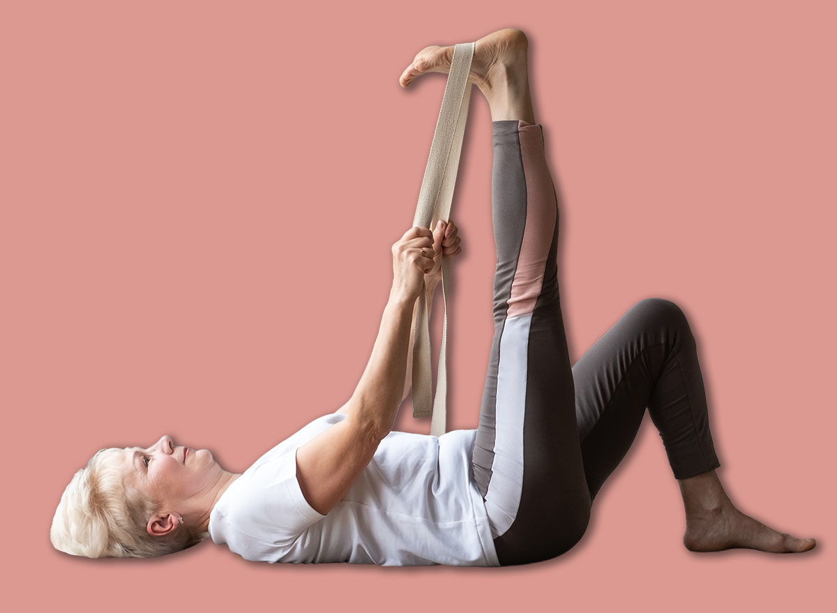 Male photographer making photos of senior woman working out, warming up using yogic belt, lying in yoga Supta Padangushthasana.