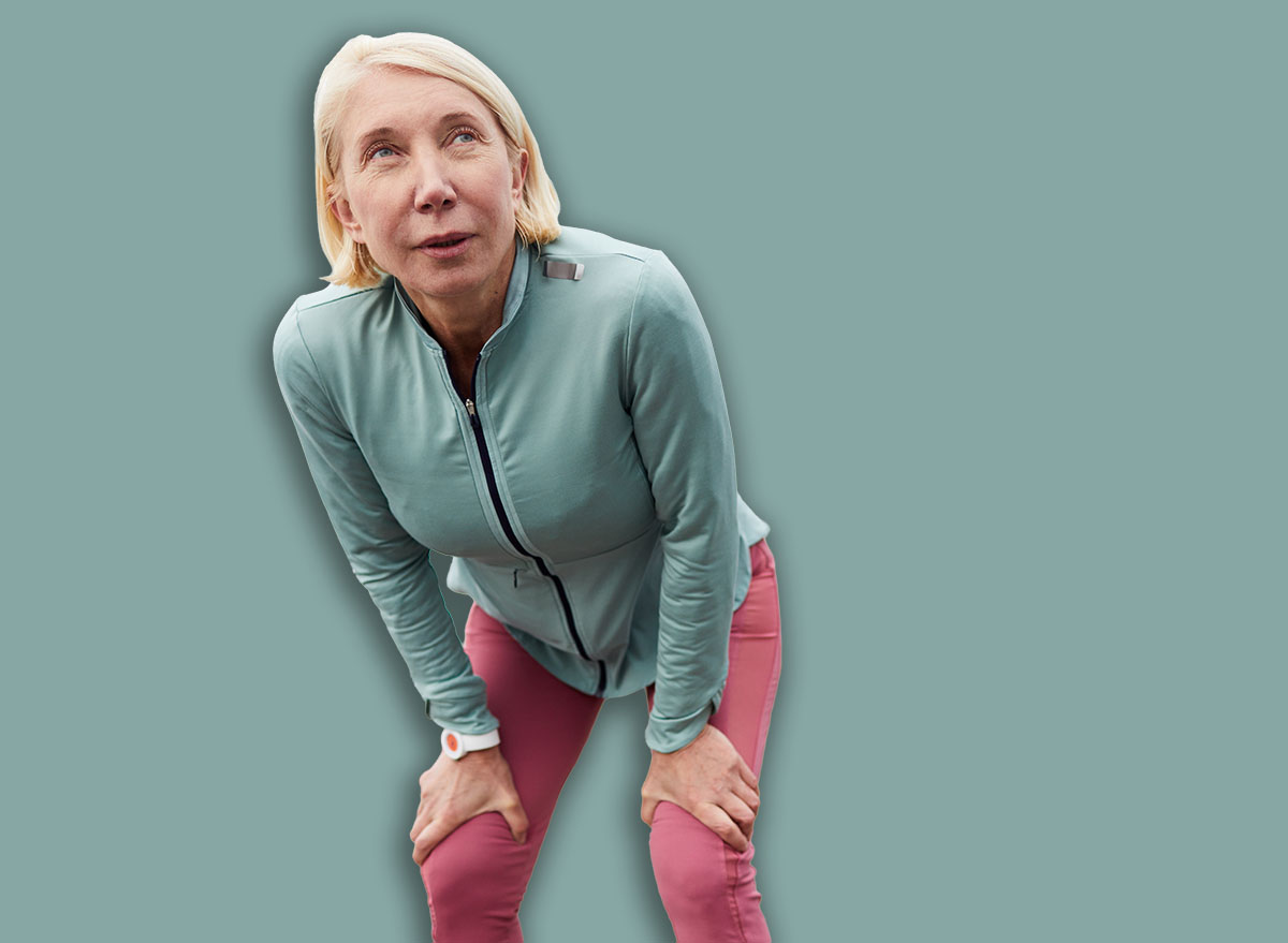 Tired mature sportswoman leaning on bent knees while standing on city bridge after jogging workout in urban environment