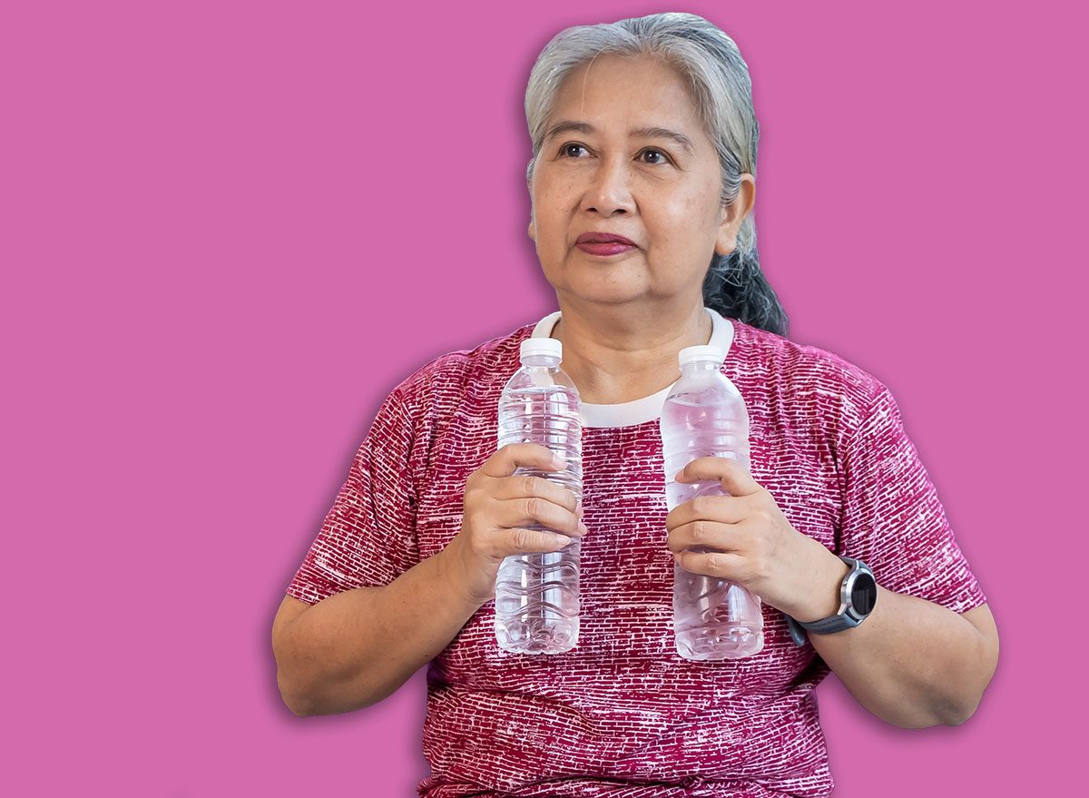 Portrait senior woman exercise with sitting on yoga mat, holding two drink water bottles on chest instead of dumbbells, home workout concept