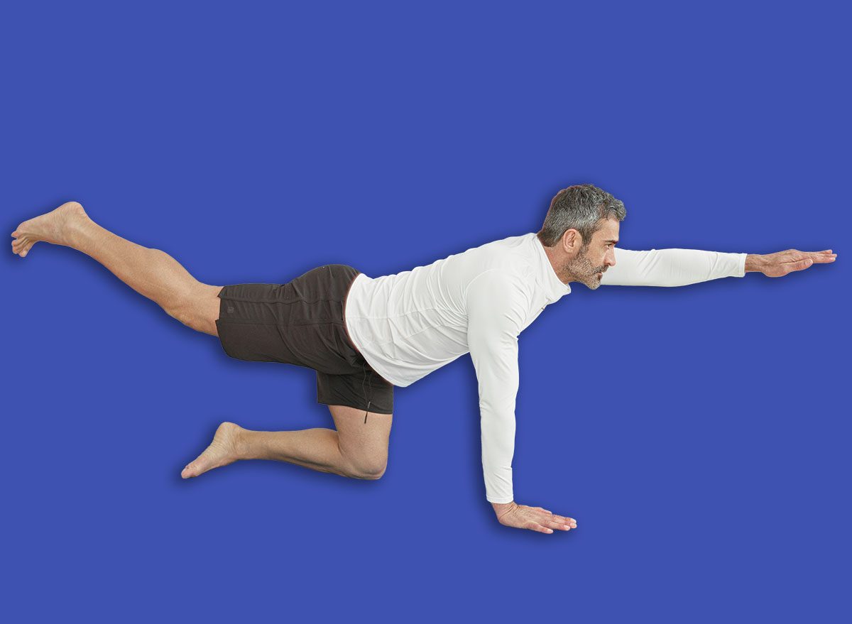 Man balancing on a blue yoga mat in studio, extending right arm and left leg in a bird dog exercise; serenity balance strength.
