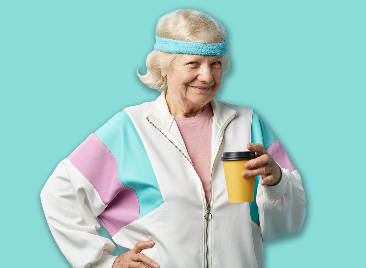 Mature female jogger in white sports suit taking a rest after morning running, holding cold drink in hands and looking at camera with cheerful look, isolated over blue background.