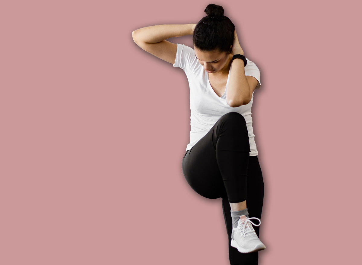 A young woman performs an abdominal crunch exercise while standing in her living room, focusing on her fitness routine