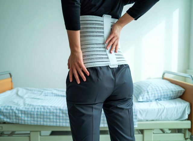 A person wearing a supportive back brace stands in a hospital room, holding their lower back, indicating discomfort or pain. The scene suggests a medical context.
