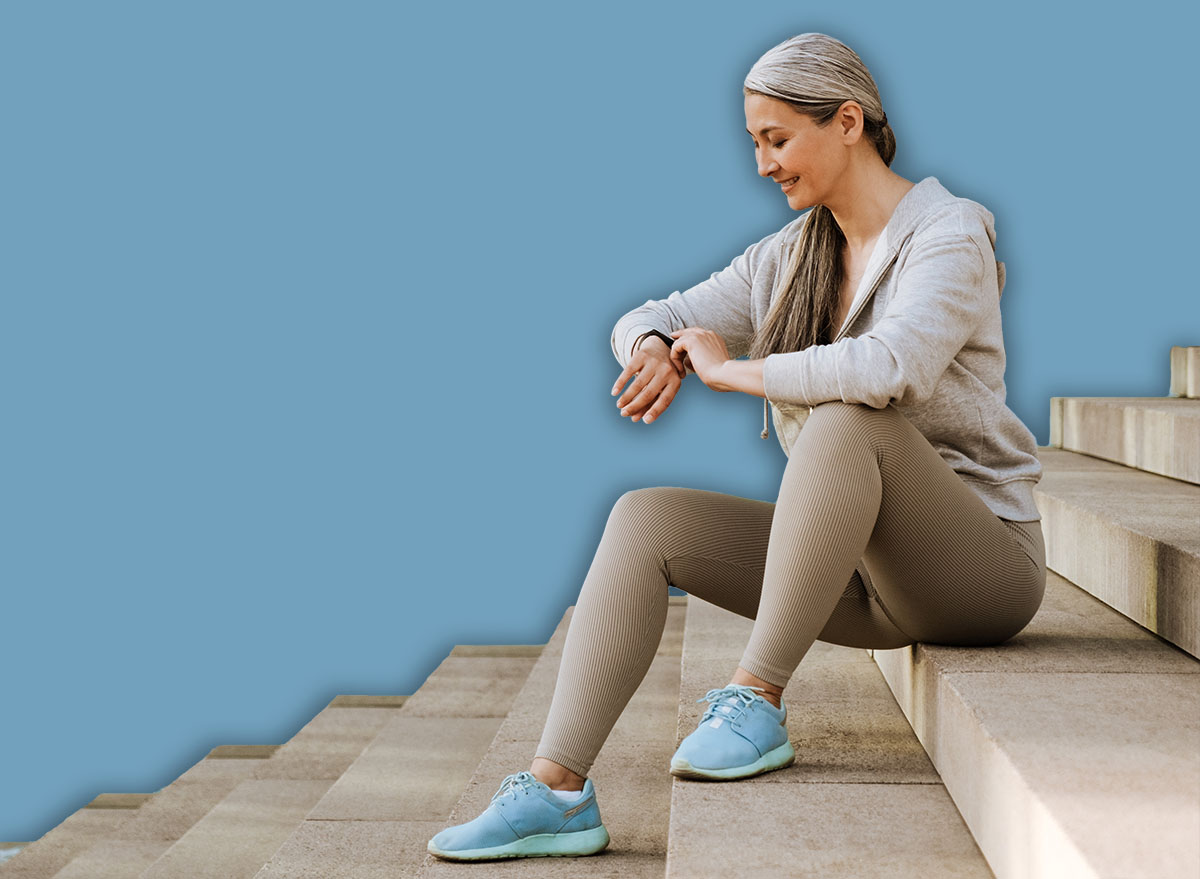 Asian mature woman in tracksuit looking ar her wristwatch and sitting on stairs during workout outdoors