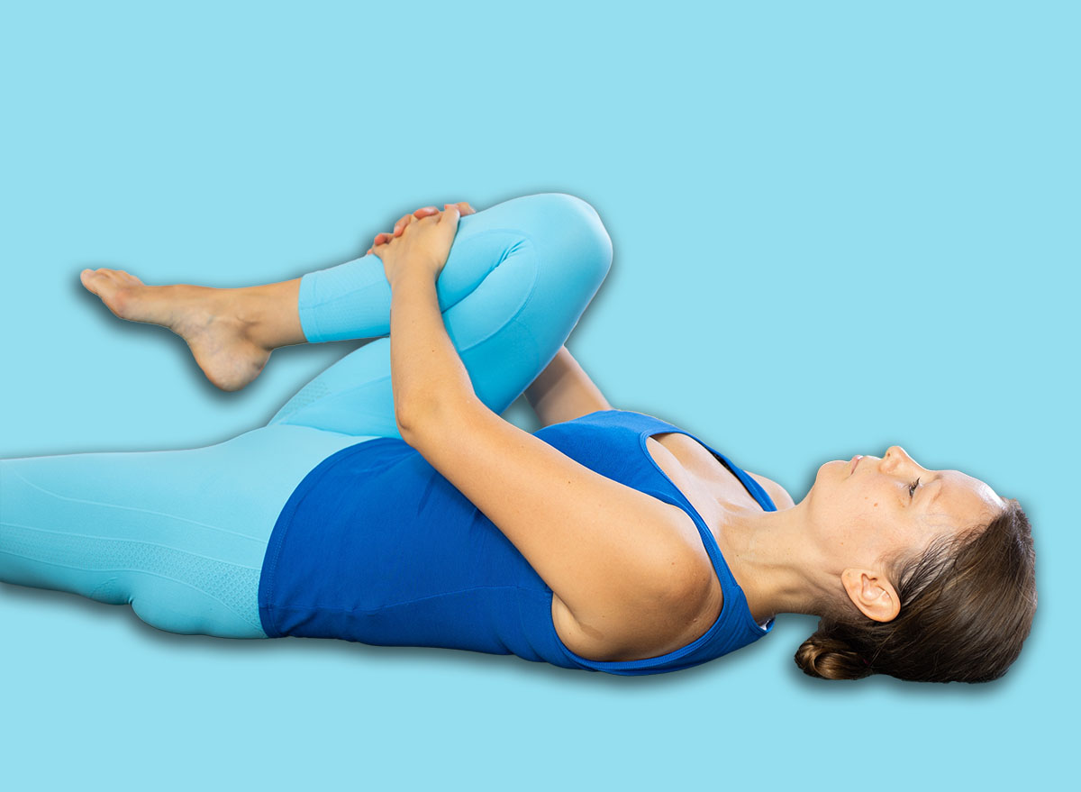 Calm middle-aged woman practicing knee to chest pose of yoga on black rug in light fitness room